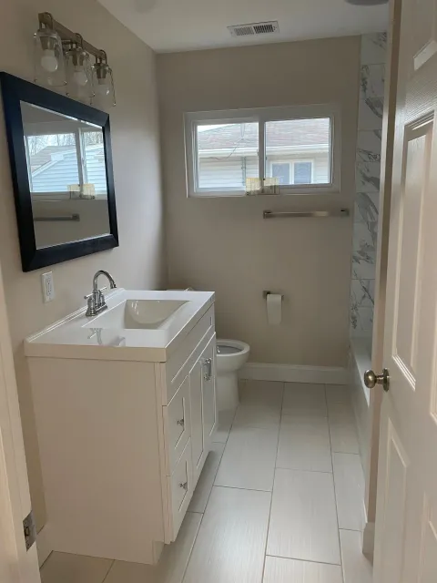 A bathroom with a sink , toilet , mirror and window.