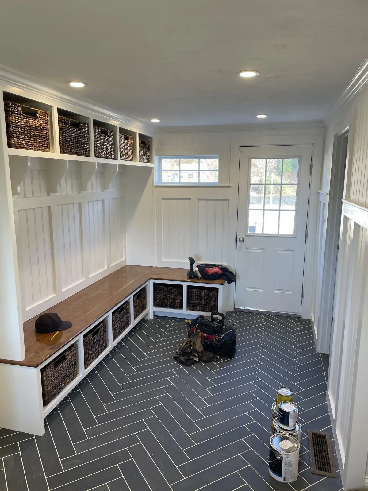 Empty entryway with white wood paneling, black herringbone tile floor, built-in bench with wicker baskets, small window, and door, tools and paint cans on the floor.