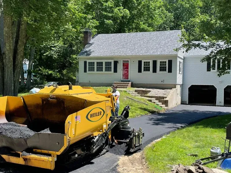 Yellow paving machine laying asphalt on a driveway in front of a white house.