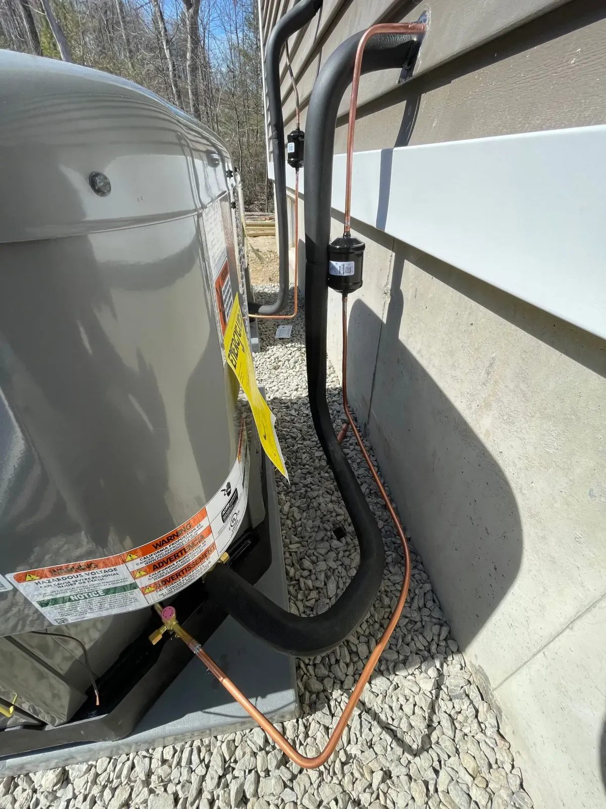 Outdoor HVAC unit beside a house, with black insulated pipes and an orange cable on gravel.