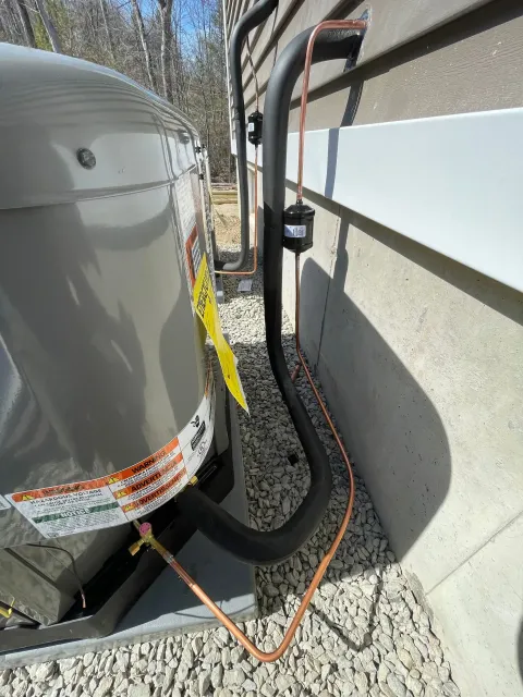 Outdoor HVAC unit beside a house, with black insulated pipes and an orange cable on gravel.