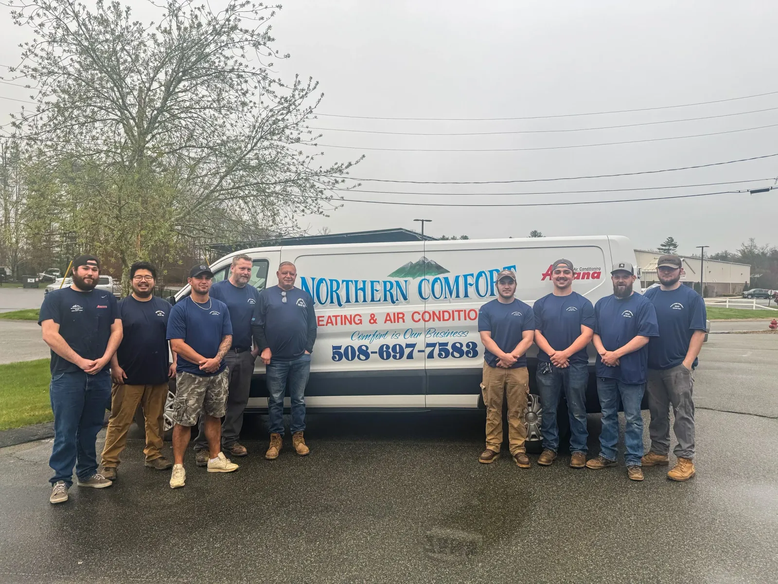 Group of nine men standing in front of a vehicle with a company logo, all wearing matching blue shirts, outdoors on a cloudy day.