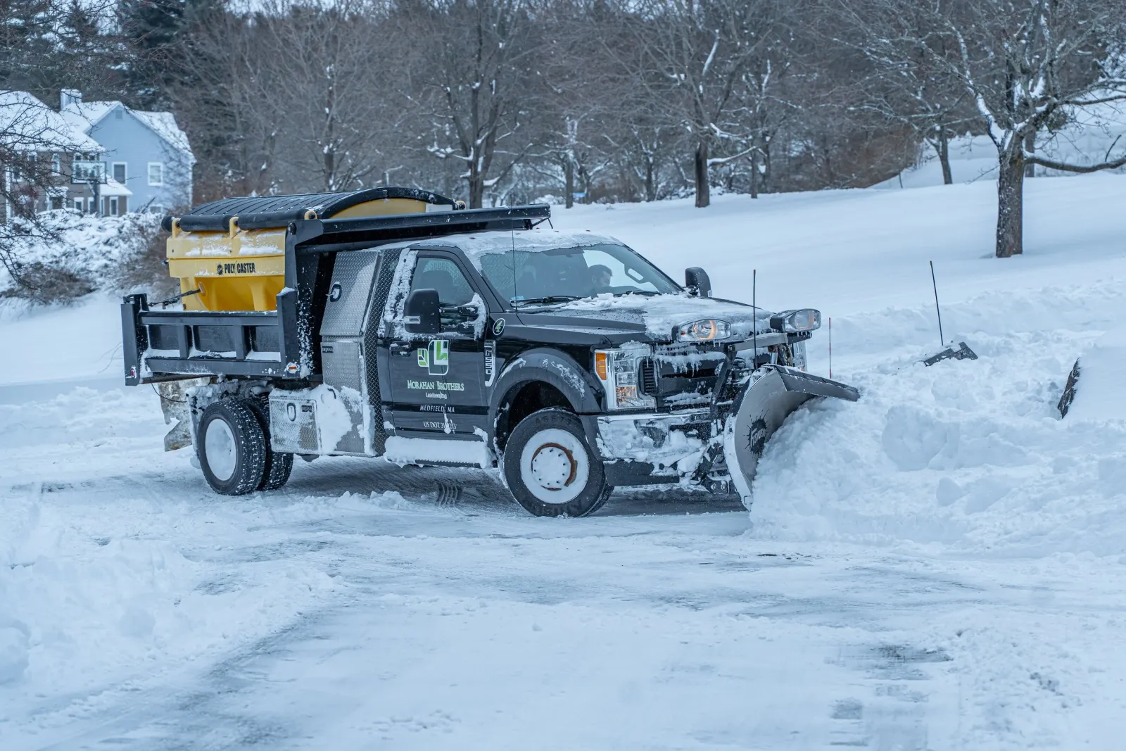 A pickup truck equipped with a snowplow clearing snow from a road in a snowy, residential area.