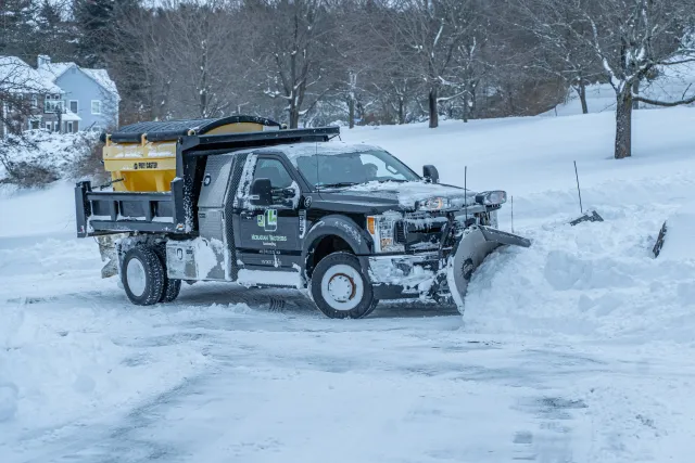 A pickup truck equipped with a snowplow clearing snow from a road in a snowy, residential area.