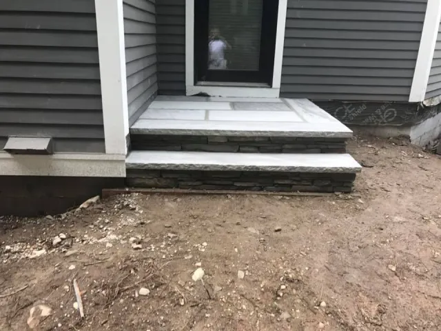 Construction site with three white stone stairs leading to a glass door, surrounded by dirt ground.