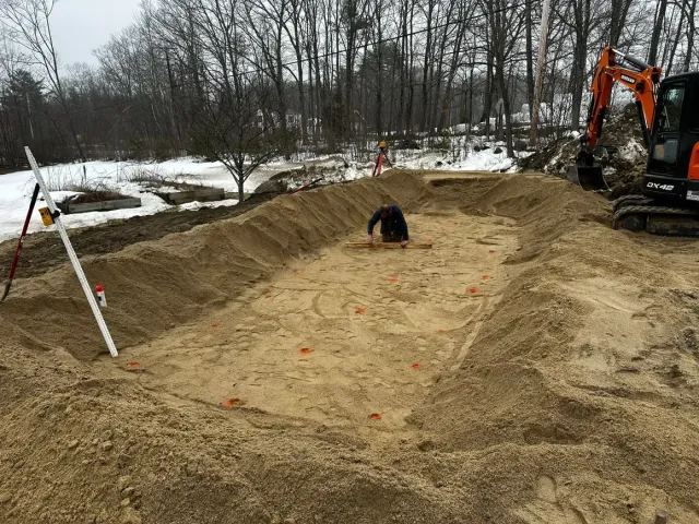 Man working inside rectangular dirt excavation, excavator nearby. Winter, trees in background.