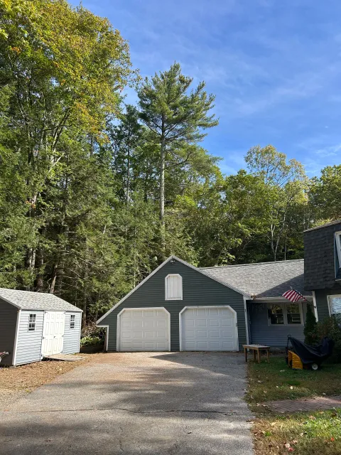 Two-car garage with white doors beside a small shed, gravel driveway, and trees under a blue sky.