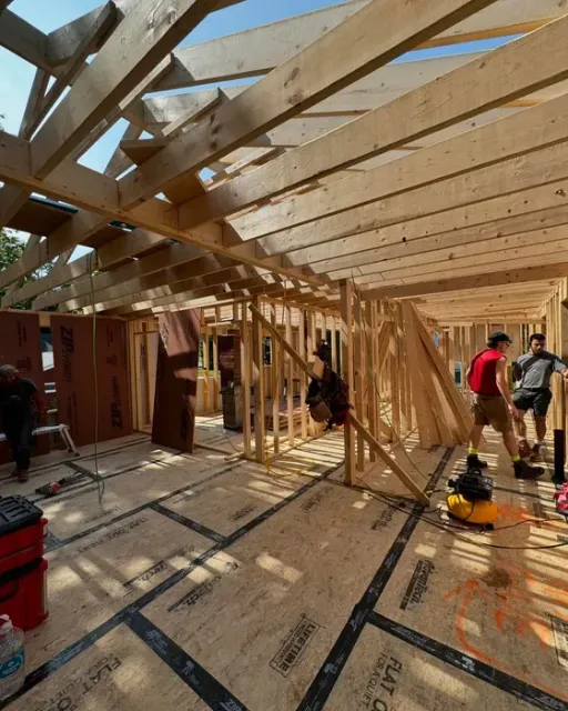 Partially framed house interior under construction, with workers and sunlight through exposed rafters.