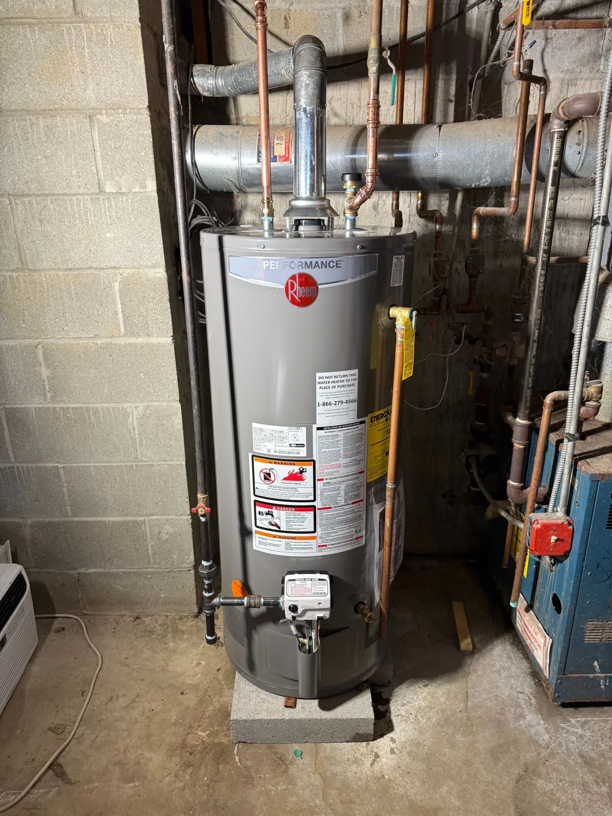 A grey Rheem gas water heater stands on a concrete block in a basement next to copper piping and a cinder block wall.