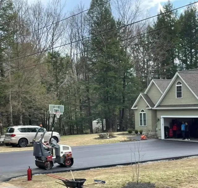 A small construction roller on a freshly paved residential driveway near a house and a car.