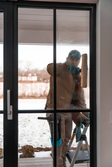 Man cleaning a glass door outside using a squeegee while standing on a step ladder in winter.