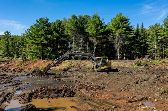 Excavator working at a muddy construction site with a dirt mound and forest backdrop