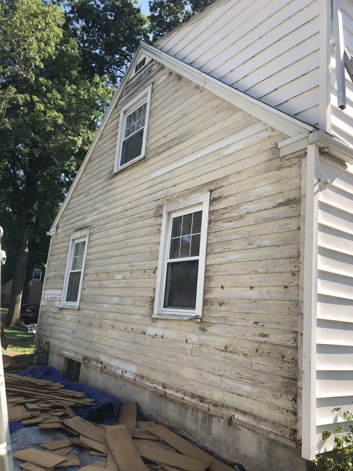 The exterior of a house with light-colored siding removed, exposing the wall sheathing, with debris on the ground.