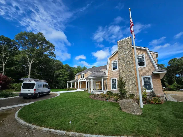 Stone house with a U.S. flag, white van, and landscaped lawn under a blue sky
