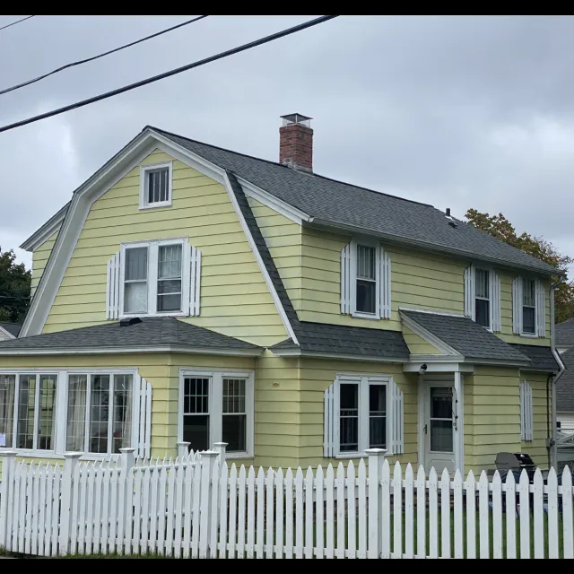 Yellow two-story house with white window shutters and a white picket fence, on an overcast day.