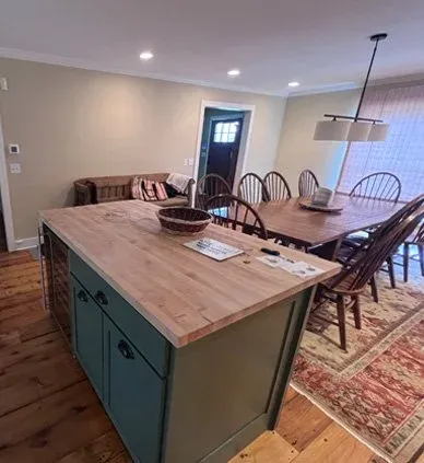 A kitchen and dining area with a wooden table, six chairs, and a wooden island with blue cabinets, a basket, and some papers on it. A rug is under the dining table, and there is a door and a window with blinds.