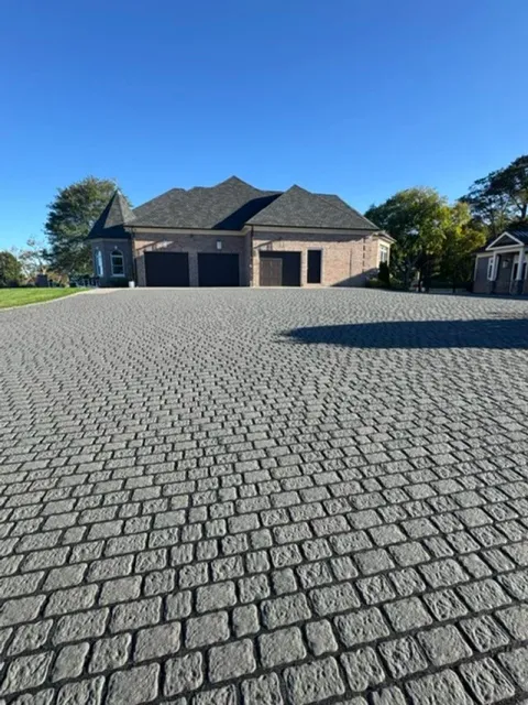 View of a large brick house with three garage doors, surrounded by a cobblestone driveway under a clear blue sky.