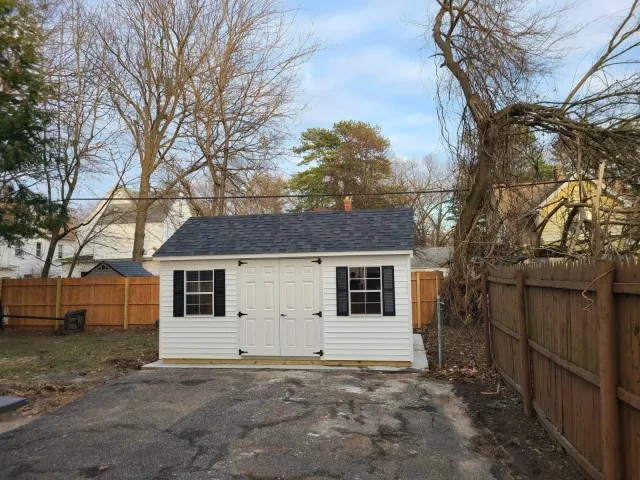 A small white shed with black shutters and double doors, situated on a concrete pad in a backyard with a wooden fence and leafless trees in the background.