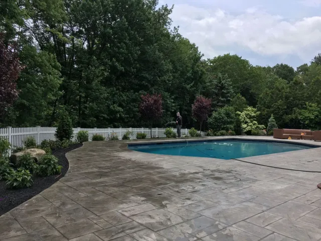 Patio beside a backyard swimming pool with trees, shrubs, and a cloudy sky in the background
