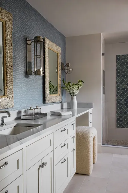 A modern bathroom with white cabinetry, a marble countertop, and a beige textured stool. There are two rectangular mirrors with ornate beige frames, a pair of sconces, and a white vase with white flowers. In the background, there's a walk-in shower w