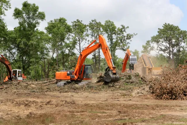 Orange excavator clearing brush in a dusty field with trees in the background.
