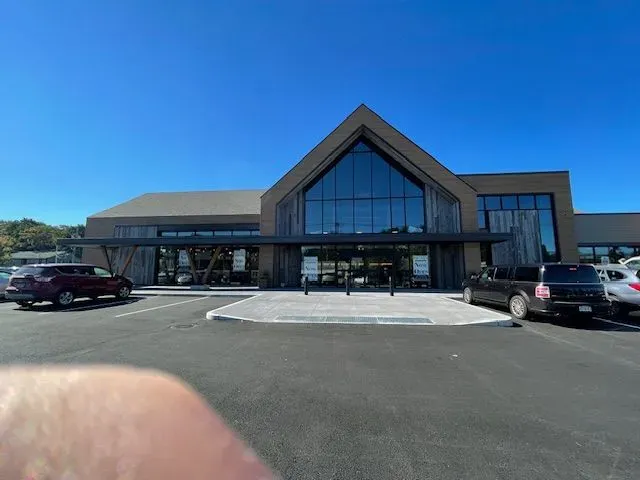 Exterior of a modern retail store with a dark wooden facade, glass entrance doors, and a parking lot.