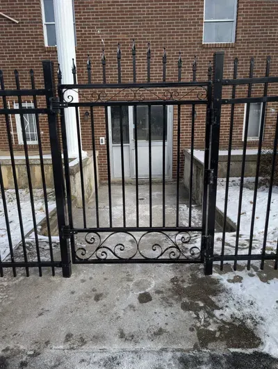 A black metal gate stands before a doorway on a snowy concrete walkway, framed by a brick building.