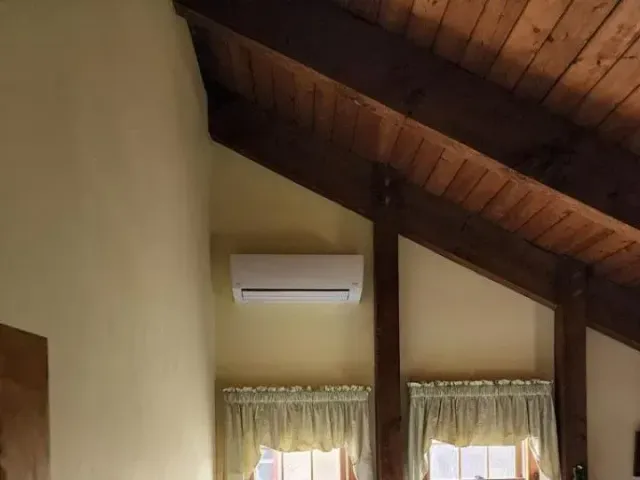 Wooden vaulted ceiling above a wall-mounted air conditioner and two curtained windows.