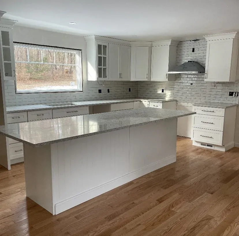 Empty kitchen with white cabinets, a large island with a light-colored granite countertop, white brick backsplash, hardwood flooring, and a window overlooking a wooded backyard.