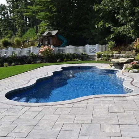 Pool with blue water and stone patio in a backyard with a playground and white picket fence.