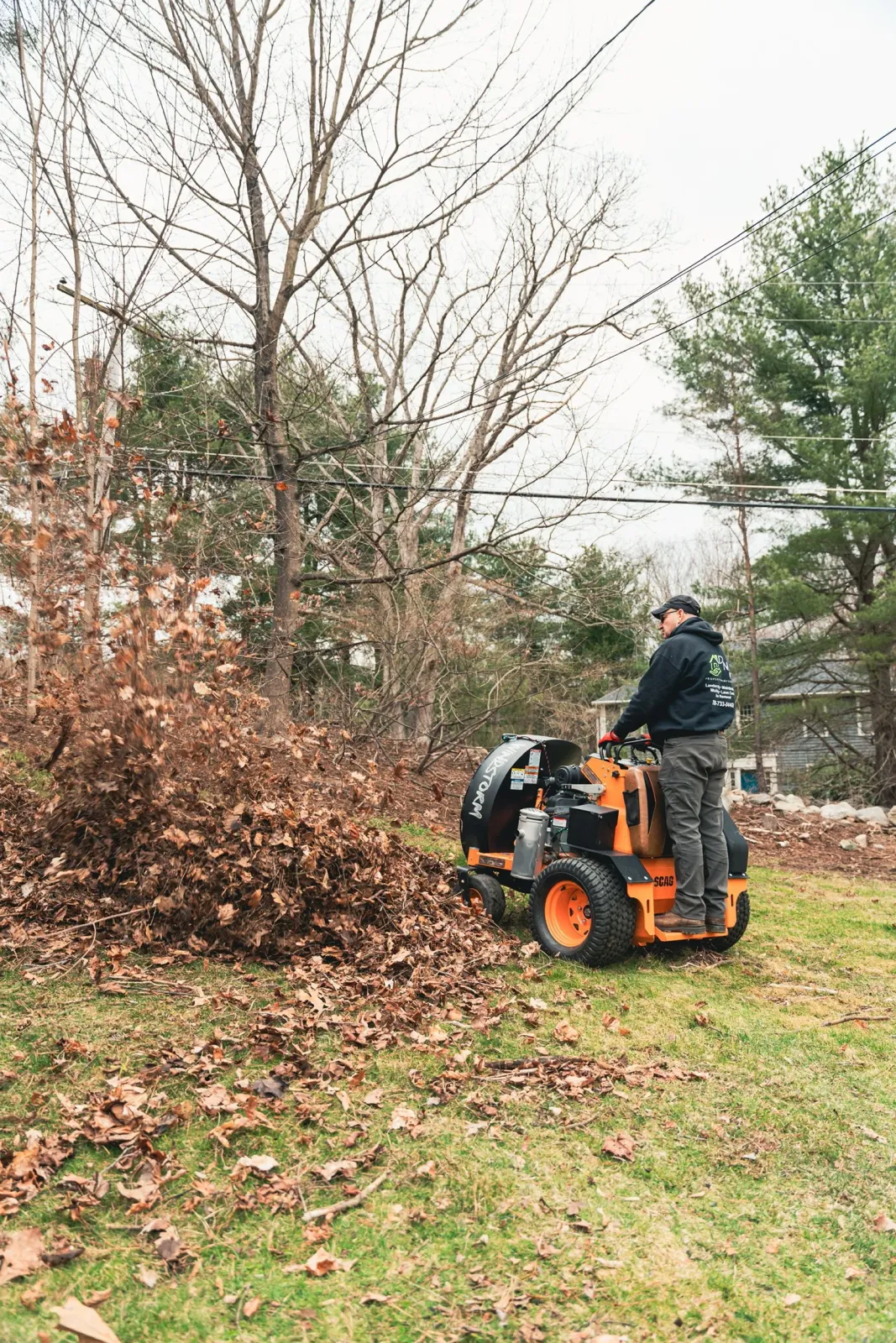 A person using a leaf blower to clear fallen leaves in a backyard yard with trees and grass.