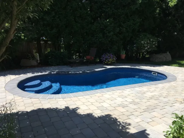 Oval-shaped pool with blue water and steps, surrounded by stone patio, lush greenery, and trees.