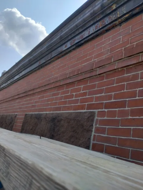 A low-angle view of a red brick wall with dark stone accents below a dark metal ledge under a blue, cloudy sky.
