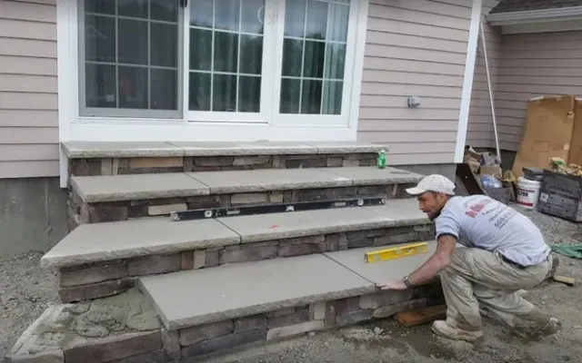 A construction worker crouches while installing stone-veneer steps on the exterior of a house, using a level for accuracy.
