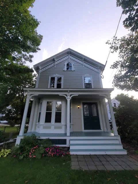 A Victorian-style house in Randolph, Massachusetts with white trim and blue exterior, featuring a front porch with steps, surrounded by grass, flowers, and trees, under a clear sky.