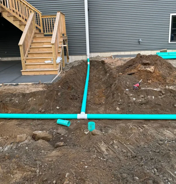 Green drainage pipes connected in a T-shape in a dirt trench next to a house with wooden stairs.
