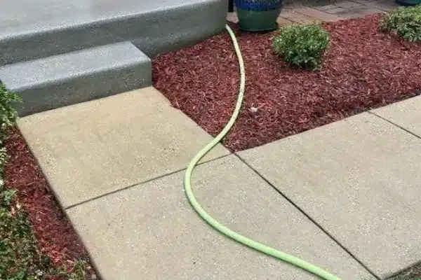 After: A green garden hose runs across a concrete walkway, bordered by red mulch and small bushes near a stone staircase.