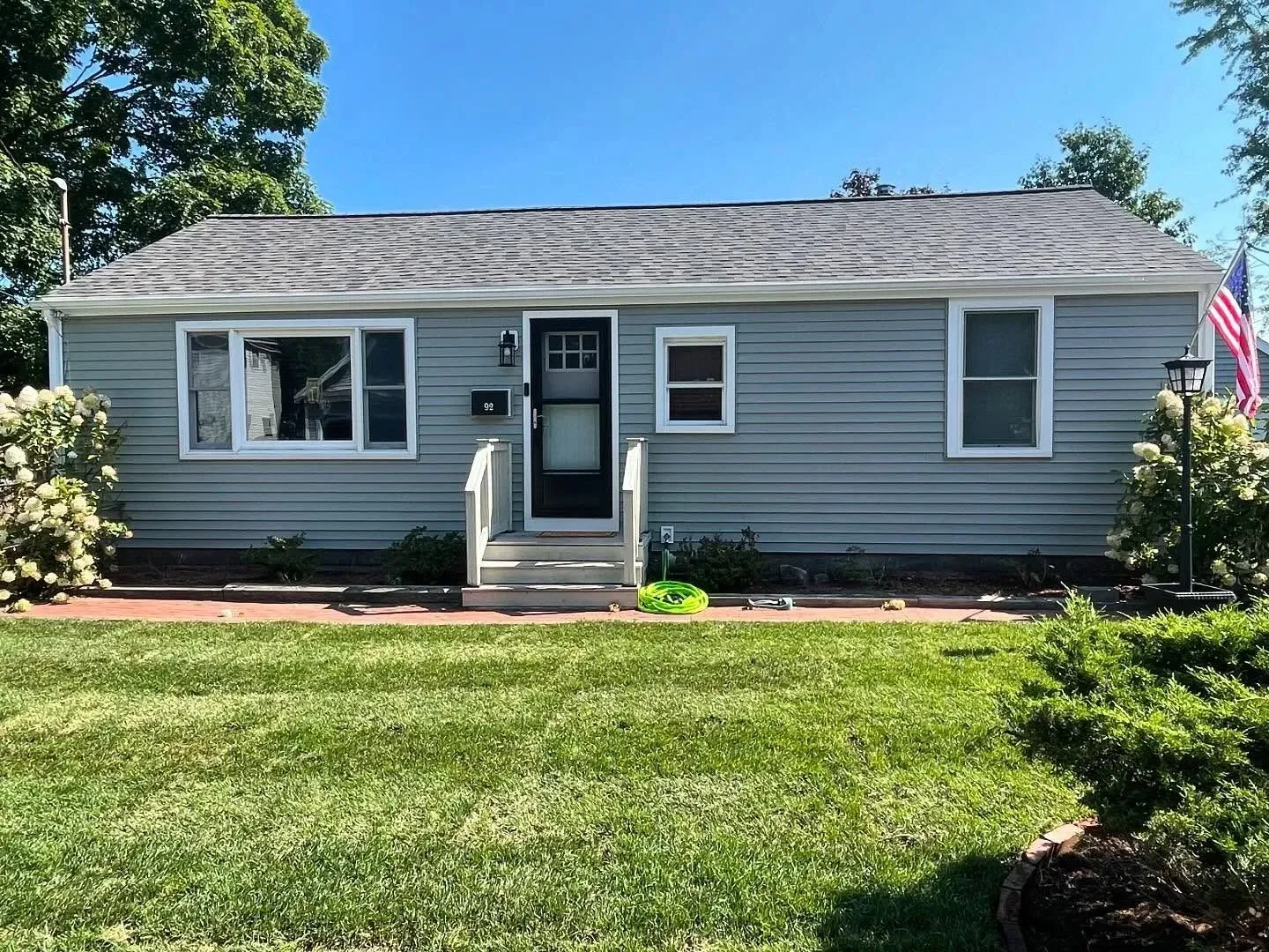 A light blue ranch-style house with gray roof shingles, a front door with a storm door, and green lawn on a sunny day.