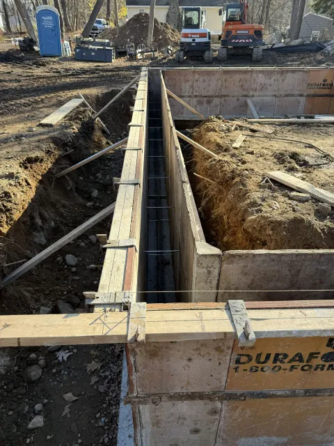 Construction site with concrete forms for foundation walls, rebar visible, excavators in background.