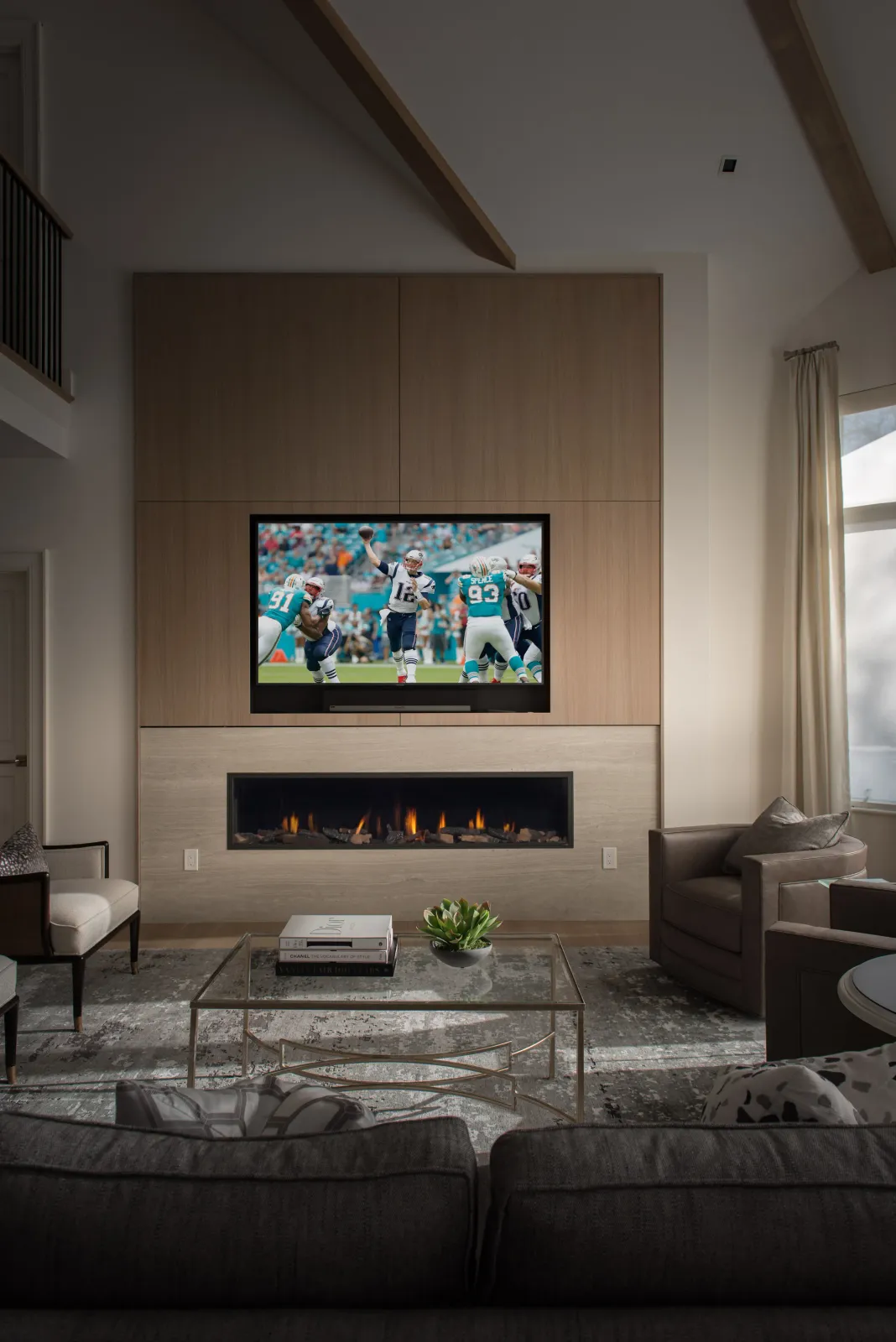 Living room with a flat-screen TV showing an American football game, above a modern fireplace, with a glass coffee table, a sofa, and armchairs in a well-lit space.