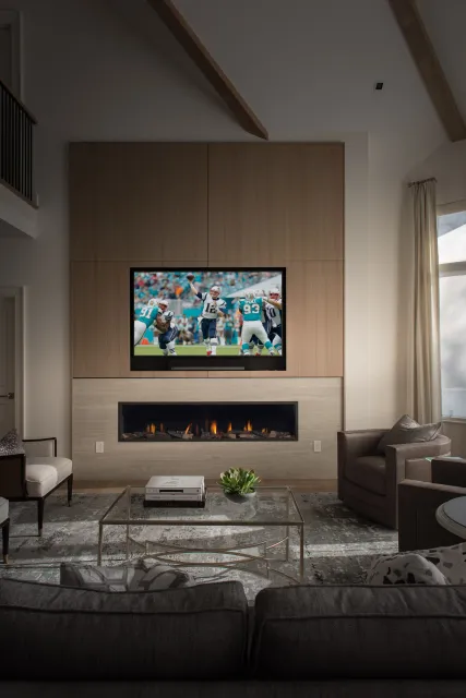 Living room with a flat-screen TV showing an American football game, above a modern fireplace, with a glass coffee table, a sofa, and armchairs in a well-lit space.