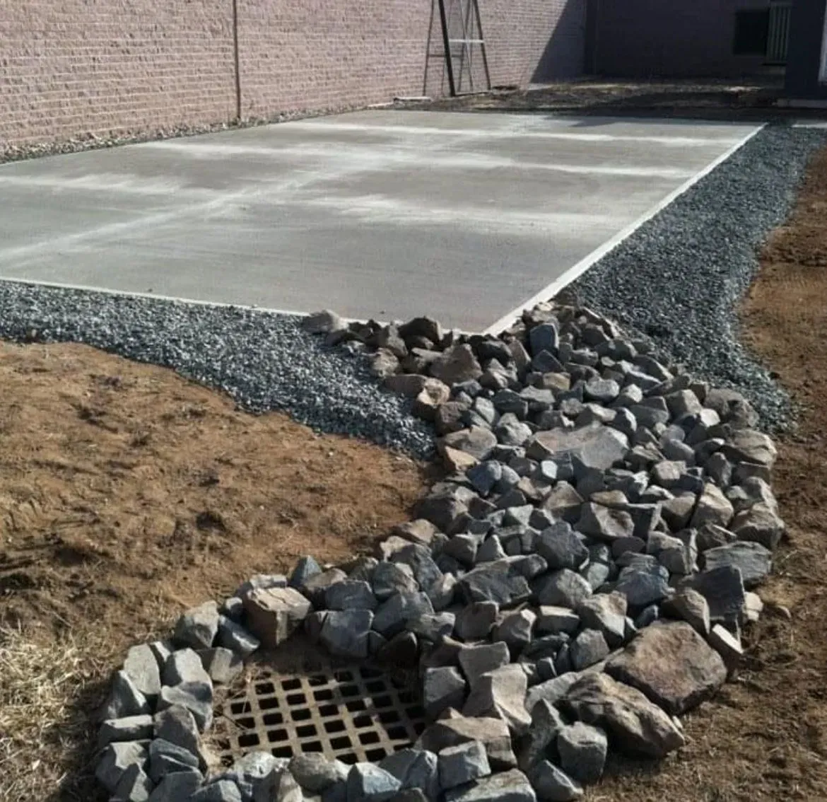 A concrete slab surrounded by gravel, featuring a rock-lined drainage basin with a metal grate in the foreground.