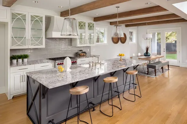 A bright, modern kitchen features a large gray island with barstools, white cabinets, and wood-beam ceilings.