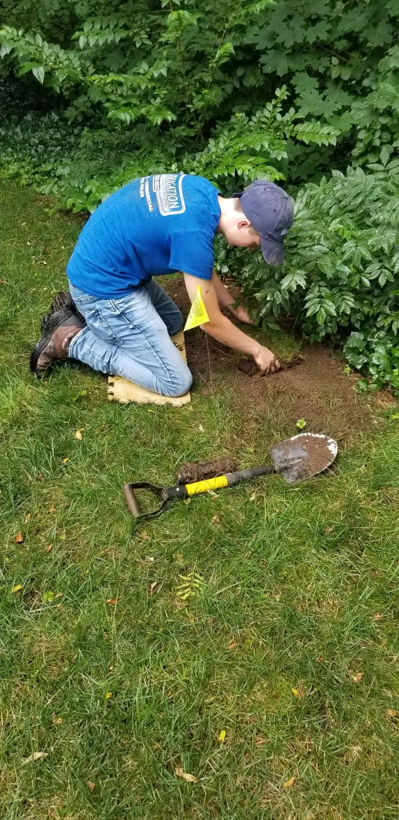 A young boy is kneeling down in the grass with a shovel.