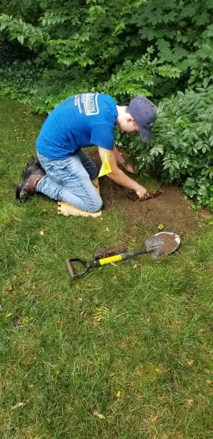 A young boy is kneeling down in the grass with a shovel.