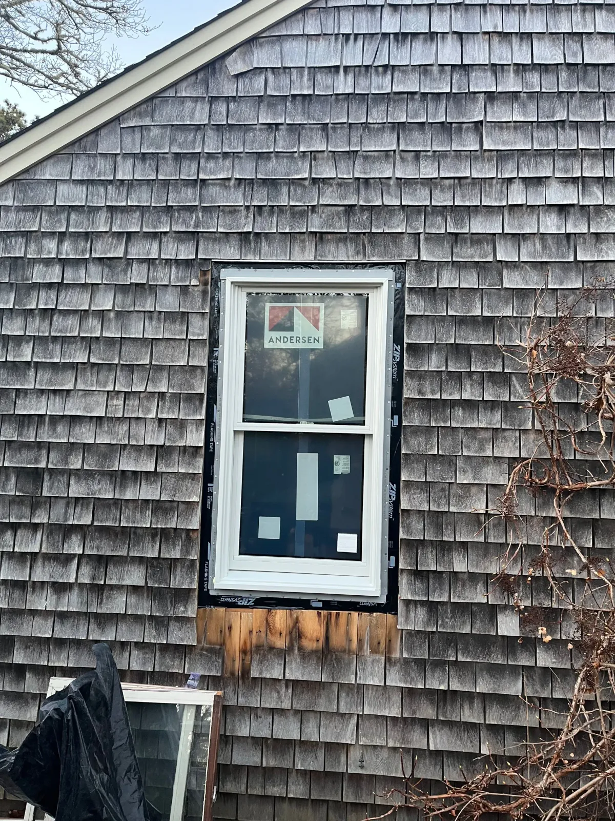 Window installation on a gray shingled house; black sealant around the white frame.