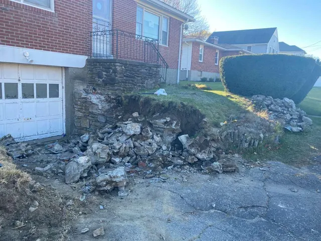Collapsed stone retaining wall beside a brick house and garage, with rubble scattered on a sloped front yard.