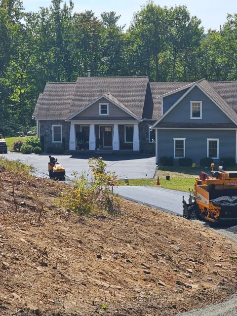 Construction equipment paving a driveway in front of a house with a grey exterior and a gabled roof.