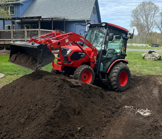 Red compact tractor with front loader moving dirt in a backyard with a house and trees in the background.