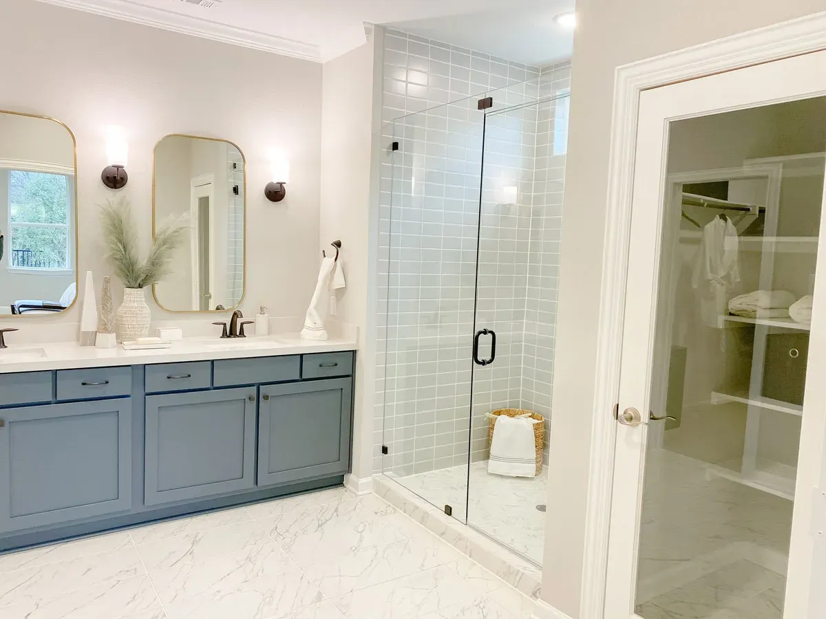 Modern bathroom with blue vanity, dual mirrors, glass shower, and walk-in closet behind glass door.